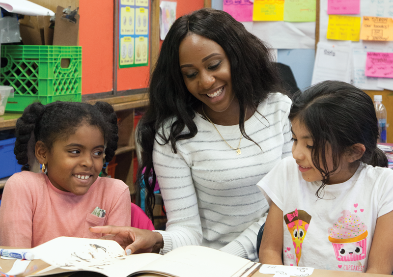 Photo of participants in the CAMBA Ready Readers program. A Black woman, a young Black girl, and a young Latina girl reading a book in a colorful classroom.