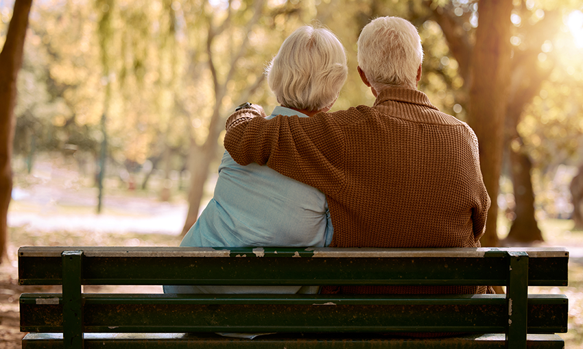 An elderly couple sits on a warm bench facing trees in a park with warm light coming through the trees.