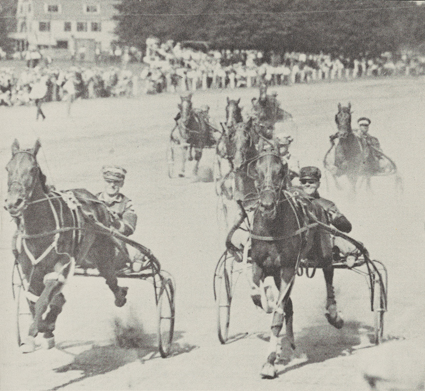 Men racing horses on a field as a crowd looks on from the sidelines.