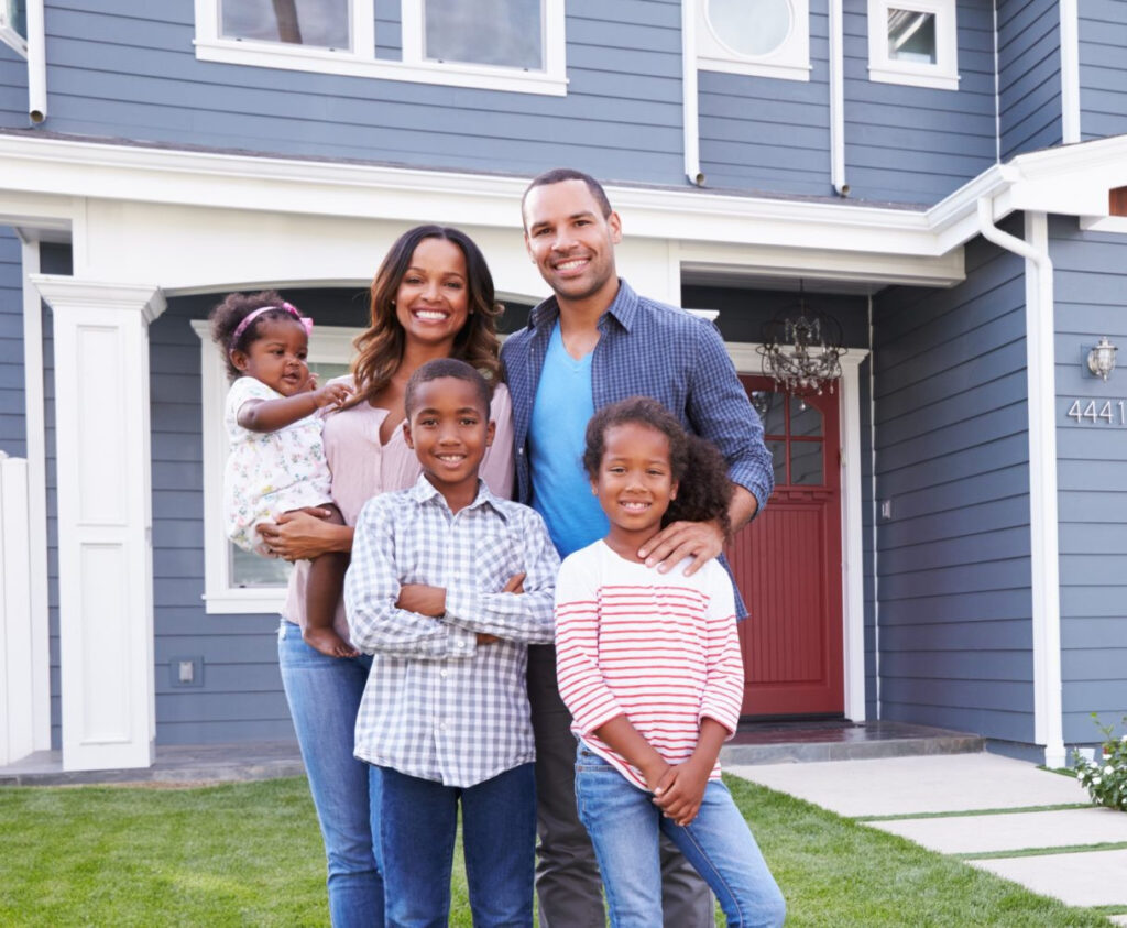 A family with three small children, a mother, and a father stand in front of a suburban house.