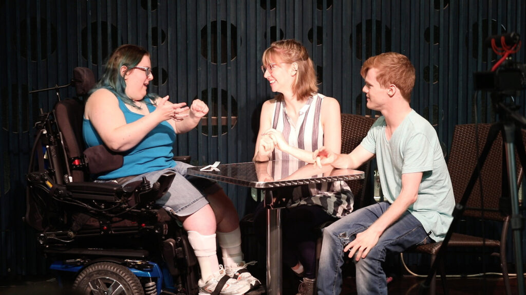 Three actors with physical disabilities sit at a small table onstage talking with each other. One actor is in a wheelchair. 