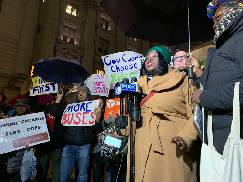 People dressed in winter coats hold signs at an evening rally. Signs read: “More buses,” “15% more service,” “More churros, less cops,” and “Gov. Cuomo, choose riders.” 