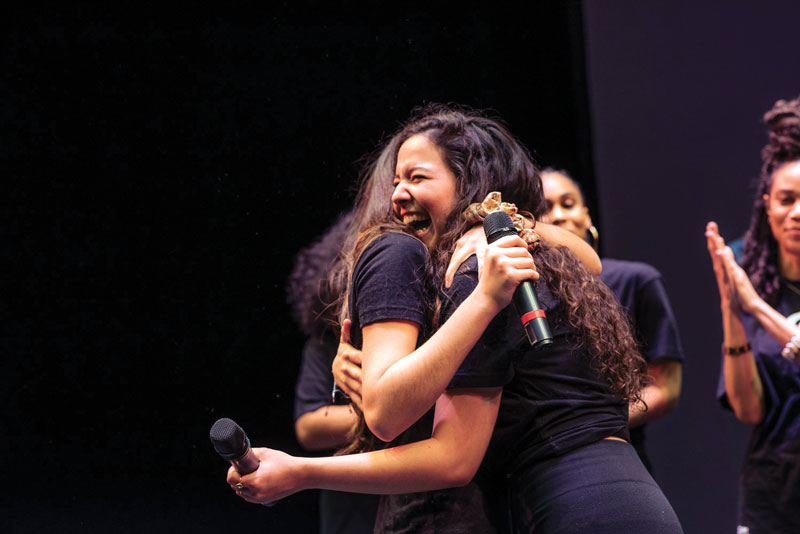 Two girls holding microphones embrace in a hug onstage  