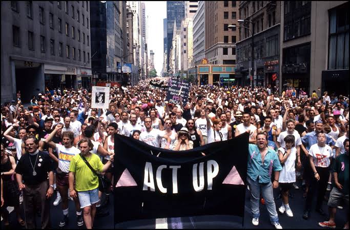 A large group of people fill the street, holding signs and a flag that states “Act Up” in 1980.