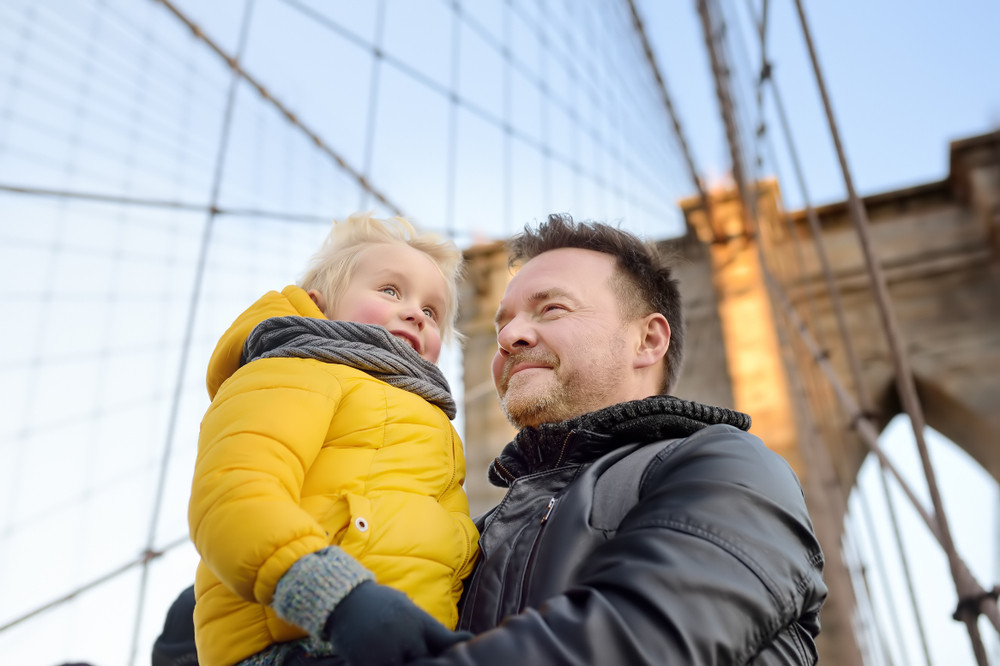 A man in a winter coat holds a toddler standing on the Brooklyn Bridge.
