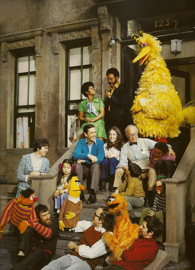 People and Muppets sitting on the steps of the Sesame Street set in the late 1900s.