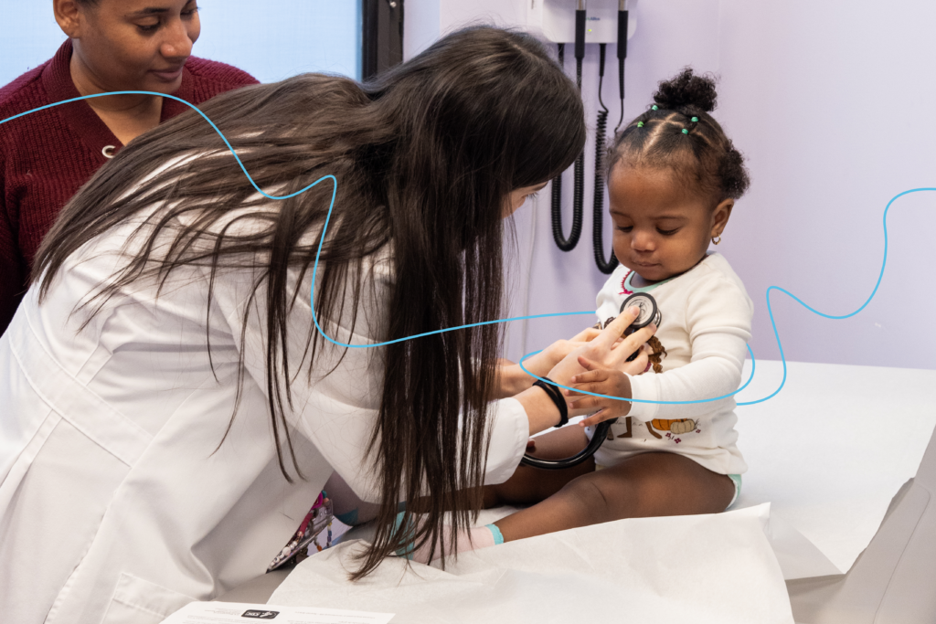A doctor listens to a baby’s heartbeat through a stethoscope while a caregiver stands by. 