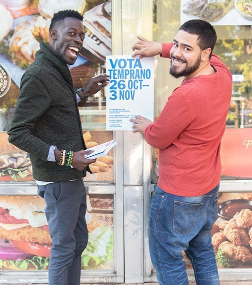 Two adults smile at the camera as they post a flyer to encourage voting. 