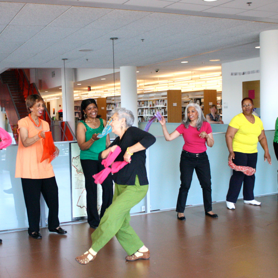 An older adult holding a bright pink scarf dances in front of five older adults who watch and smile while wearing colorful shirts and waving brightly colored scarves. 