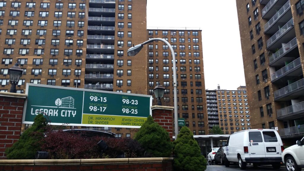 A city sign that reads, “Lefrak City” with tall buildings and vehicles in the background.