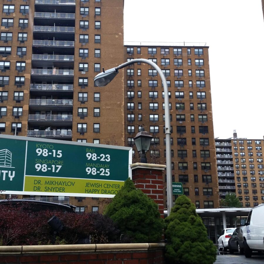 A city sign that reads, “Lefrak City” with tall buildings and vehicles in the background.