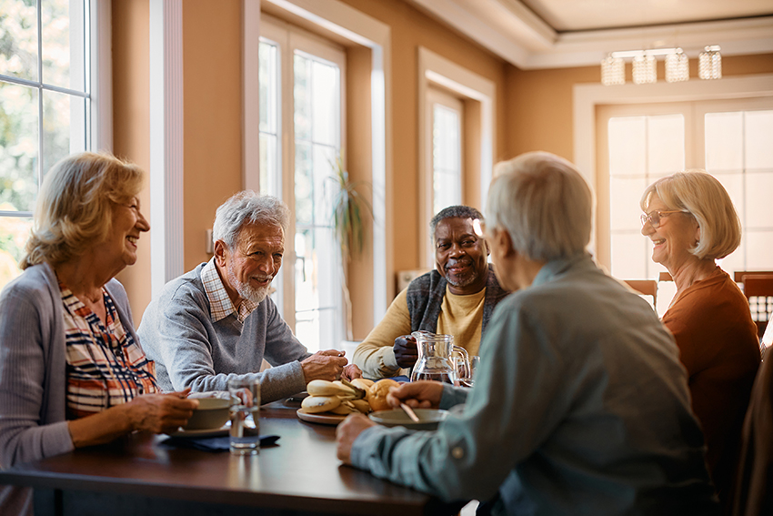 A group of seniors sitting around a table with food, talking and smiling as warm light comes in the large windows.