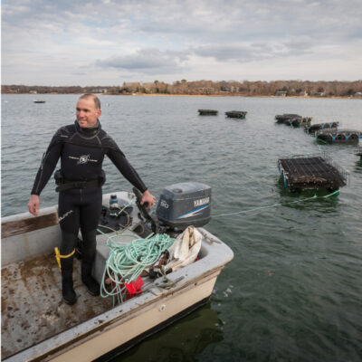 An adult wearing a wetsuit stands in an oyster boat on the Peconic Bay.