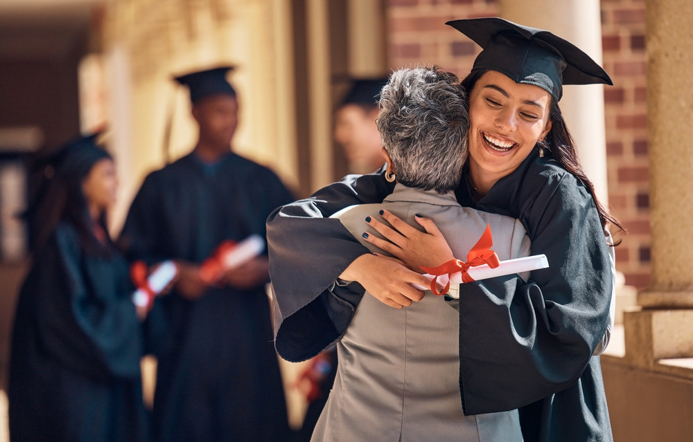 Smiling graduate wearing robe and mortarboard hat hugs an older person. 