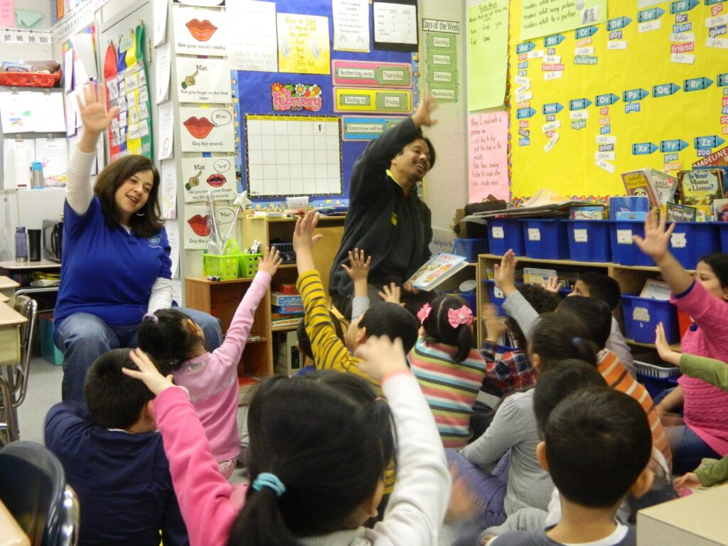 Two educators sitting on chairs in a classroom raising their hands, engaging with a group of young students sitting on the floor and raising their hands in response.