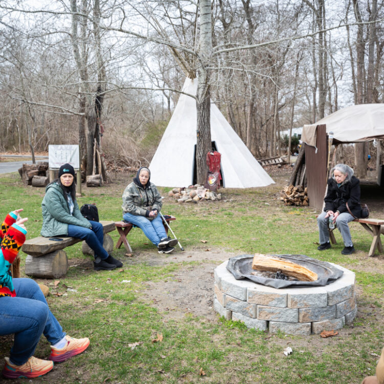 Tribal members in front of a teepee and firepit outside.