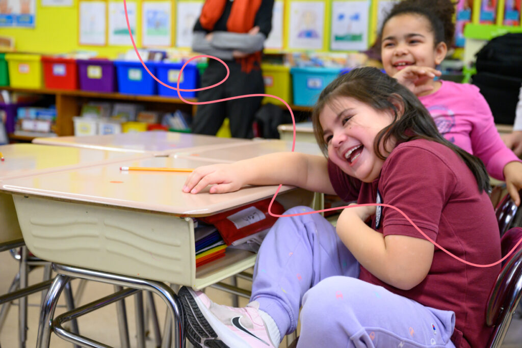 two elementary school students laugh as they sit at their desks. 