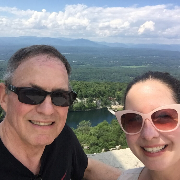 A man and a woman wearing sunglasses smile for the camera overlooking the Mohonk Preserve in upstate New York. 